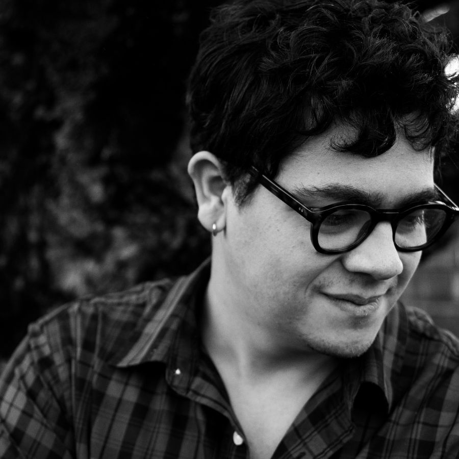 A black and white portrait of poet Gabriel Reed. Reed is smiling and looking down and away from the camera.