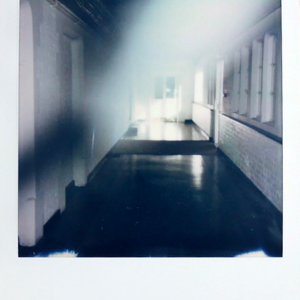 A Polaroid photograph of a long corridor with a dark floor and white walls. There is sunlight at the end of the corridor. A blur is obscuring the photo's foreground.